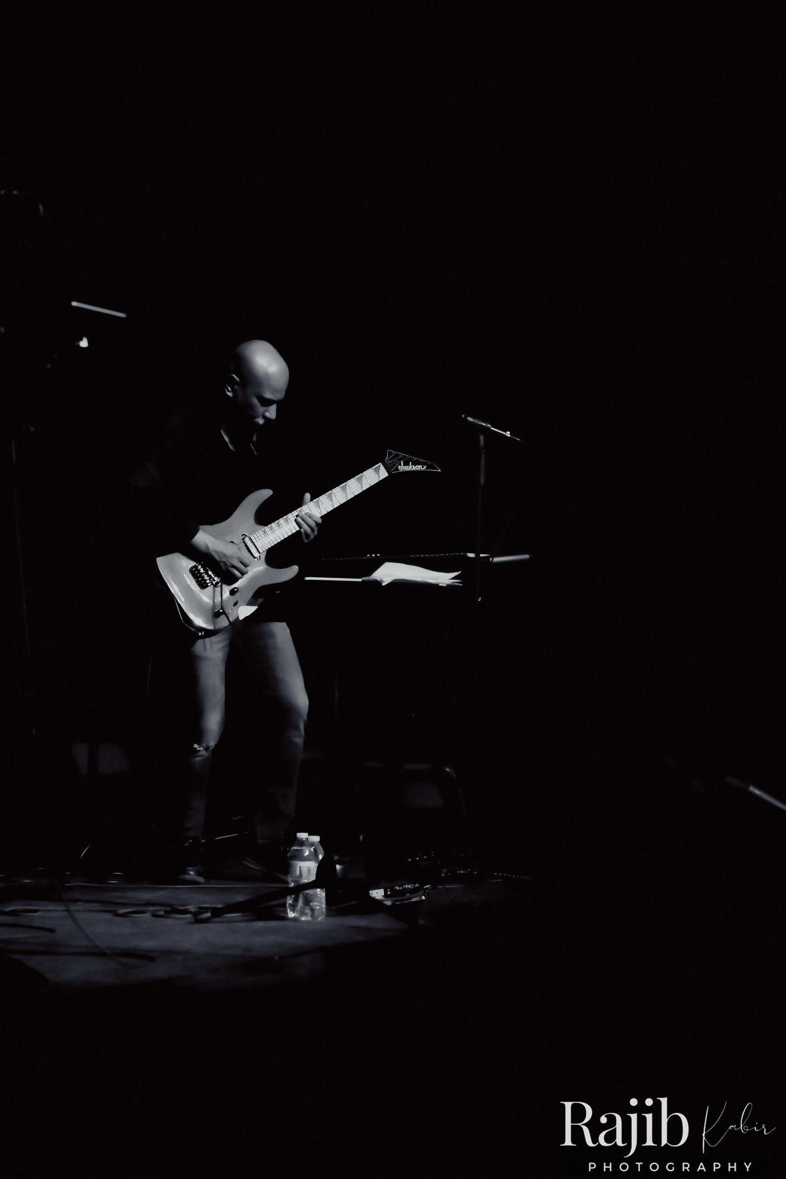 A musician playing an electric guitar on stage, illuminated by stage lights in a monochrome setting.
