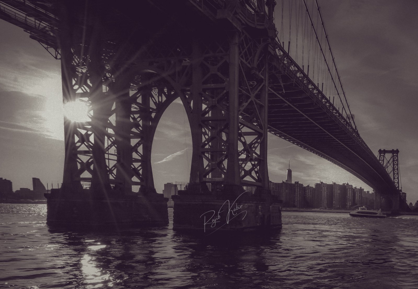 Williamsburg Bridge in New York City, captured by Rajib Kabir in black and white, with the skyline and sun peeking through its steel framework.